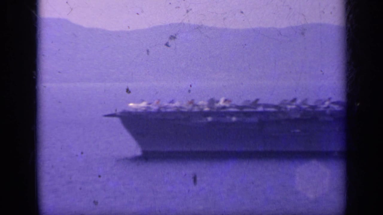 1962 Italy-associated home-movie frame showing the carrier bow and flight deck aircraft in soft purple-tinted 8mm footage.