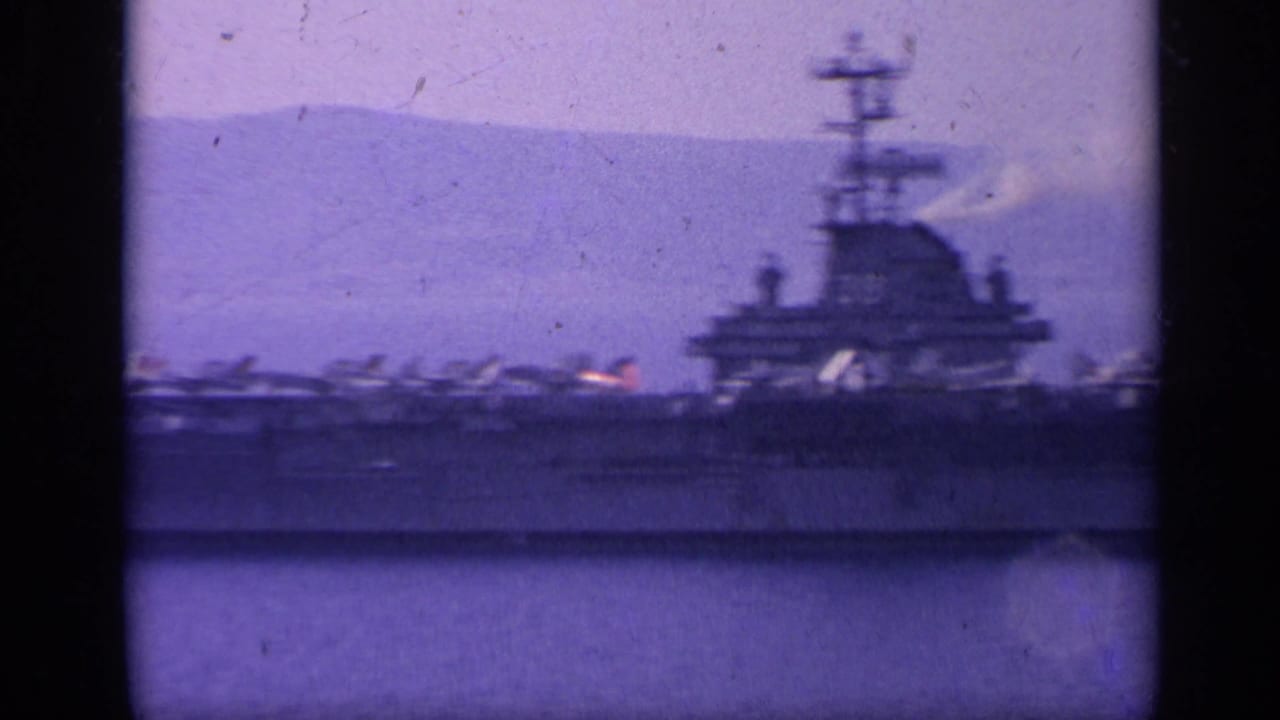 Distant side view of a U.S. Navy carrier in Italy-associated 1962 8mm footage, with island structure and deck aircraft visible.