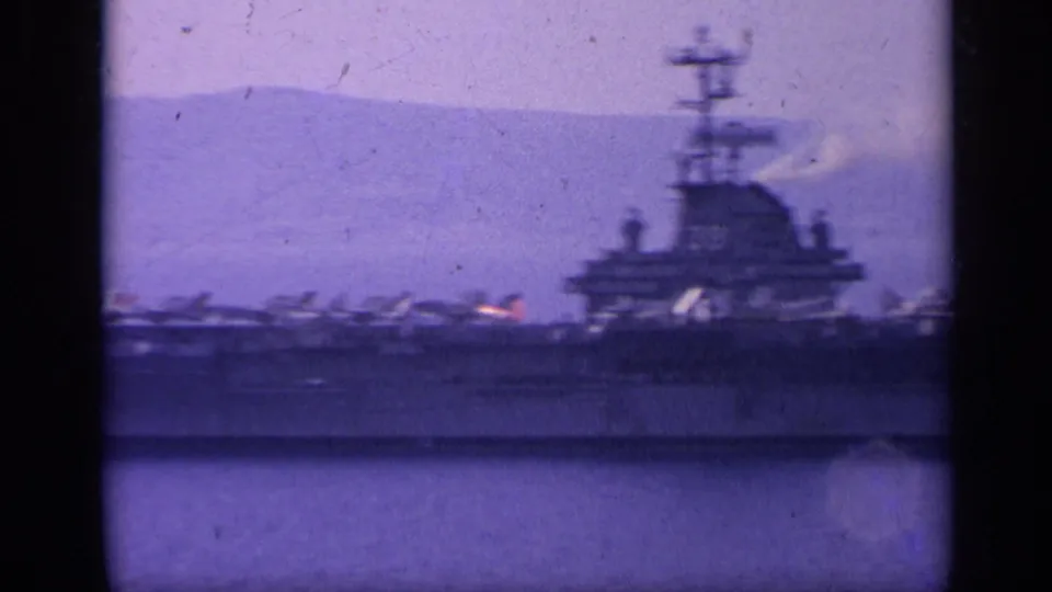 Distant side view of a U.S. Navy carrier in Italy-associated 1962 8mm footage, with island structure and deck aircraft visible.