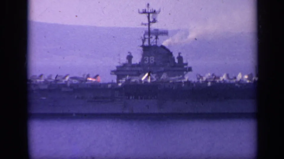 1962 Italy 8mm frame showing a U.S. Navy aircraft carrier marked 38, with aircraft on deck and mountains beyond the water.