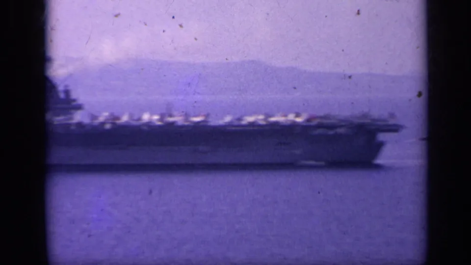 A distant broadside view of the U.S. Navy carrier off Italy in 1962, with deck aircraft and a hazy mountain horizon.