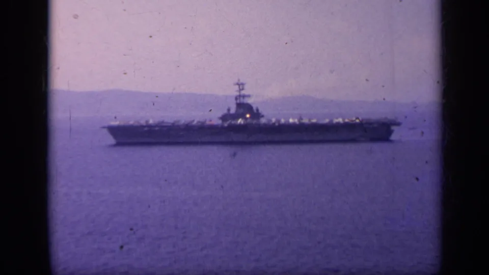 Wide 1962 Italy frame of a U.S. Navy aircraft carrier on calm open water, seen from a distance with hazy mountains behind.