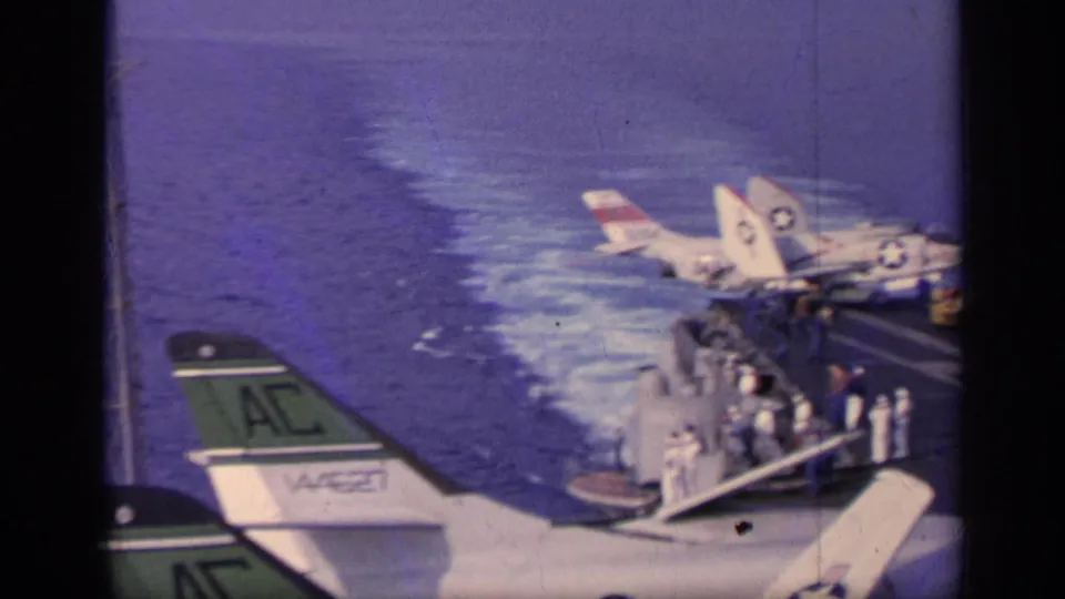 1962 Italy 8mm frame showing U.S. Navy jets parked on a carrier deck, with crew gathered near railings and the ship wake alongside.