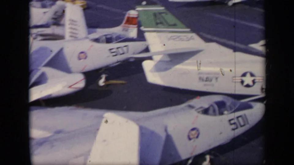 Close 1962 carrier-deck view of a white Navy jet marked 507 with other aircraft and U.S. insignia nearby.