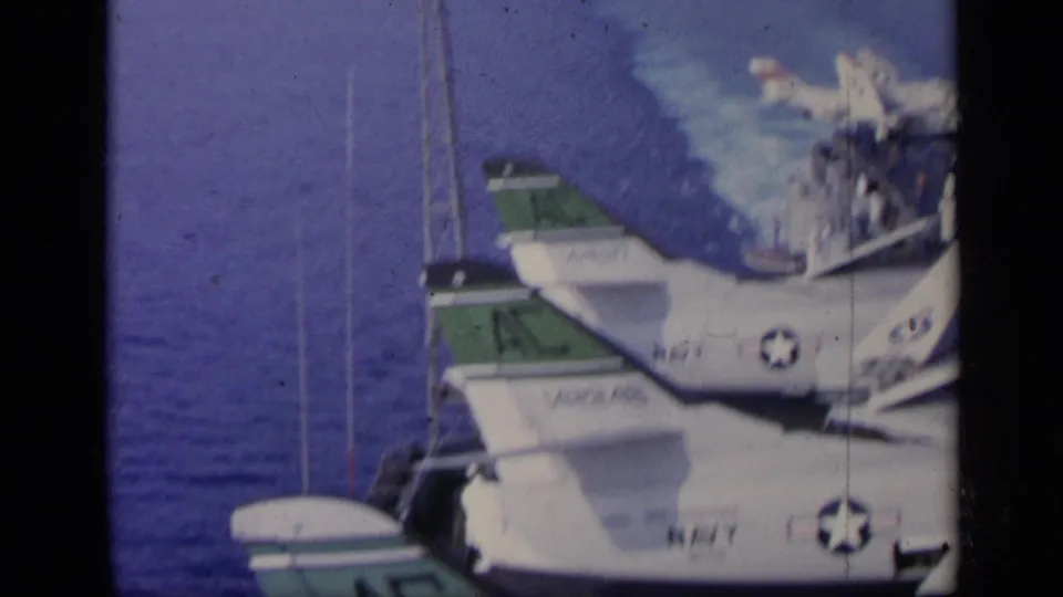 1962 Italy home-movie frame showing white Navy aircraft with green tail sections on a carrier deck above open water.