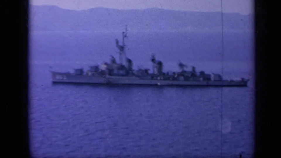 A gray U.S. Navy-style warship appears in profile on calm blue water near a hazy mountainous coastline.