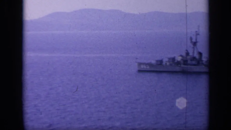 A gray naval vessel is partly framed near the right edge, with calm water and hazy coastal mountains behind it.