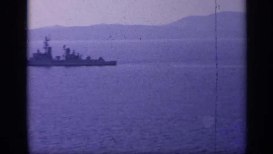 A distant gray warship sits low on calm blue water, framed by hazy mountains and visible 8mm film edges.