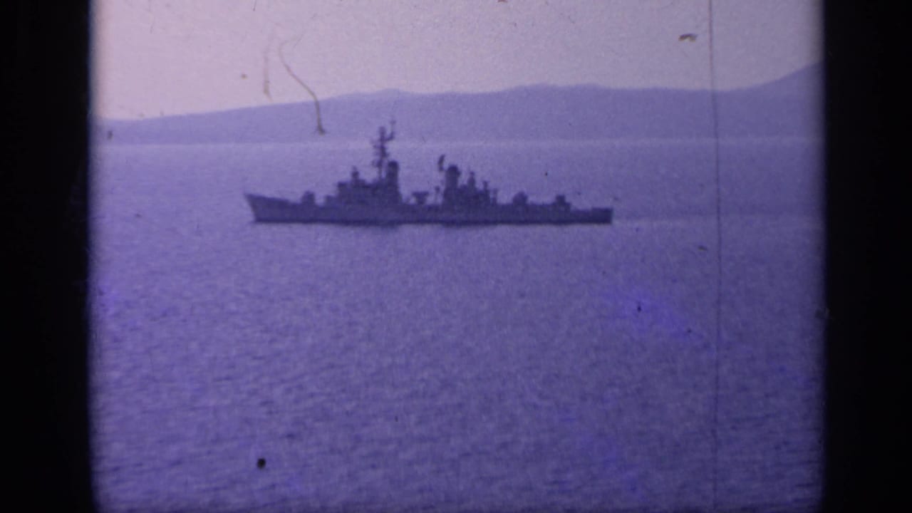Naval warship on calm water with hazy mountains behind it.