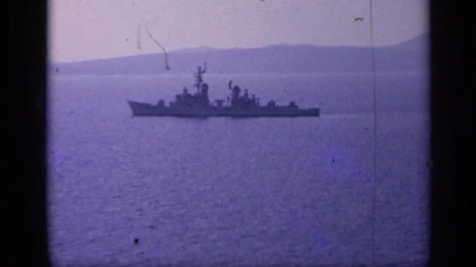 A distant naval warship crosses calm water near a hazy mountainous Italian coastline in 1962 8mm footage.