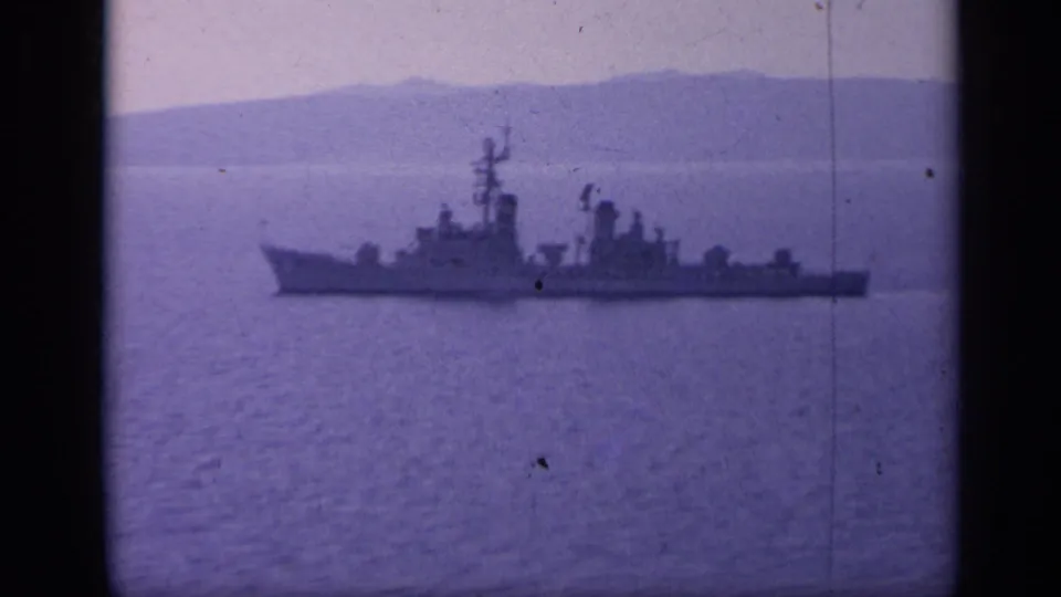 The warship appears larger in frame as it moves across blue-purple water with a distant Italian mountain coast behind it.