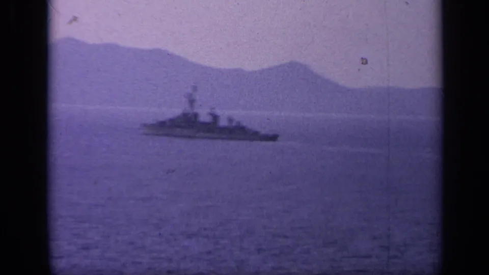 A naval ship recedes toward the left side of the frame, set against hazy mountains and calm Italian coastal water.