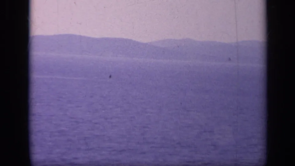 The final frame holds on calm blue-purple water and a hazy mountainous Italian coast after the warship exits view.
