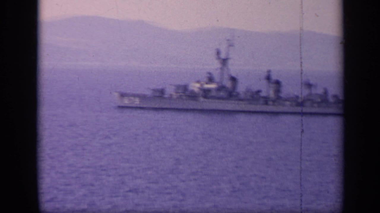 A distant naval vessel crosses water near a hazy mountain coastline.