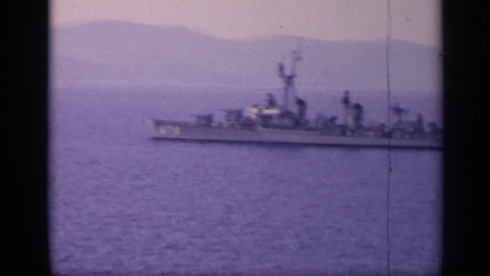 1962 Italy 8mm frame with a distant gray naval vessel moving across purple-tinted coastal water below hazy mountains.