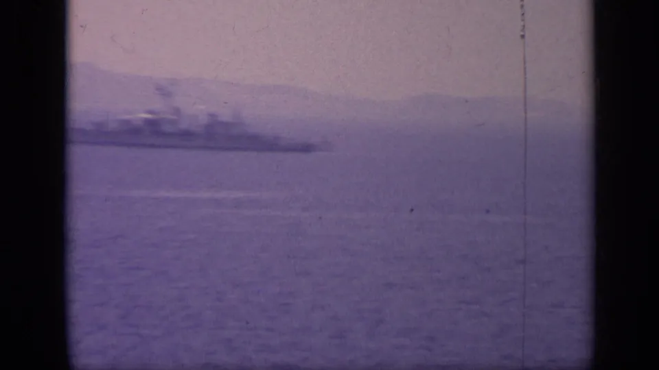 1962 Italy 8mm frame with a blurred gray naval vessel receding across coastal water beneath a hazy sky and mountains.