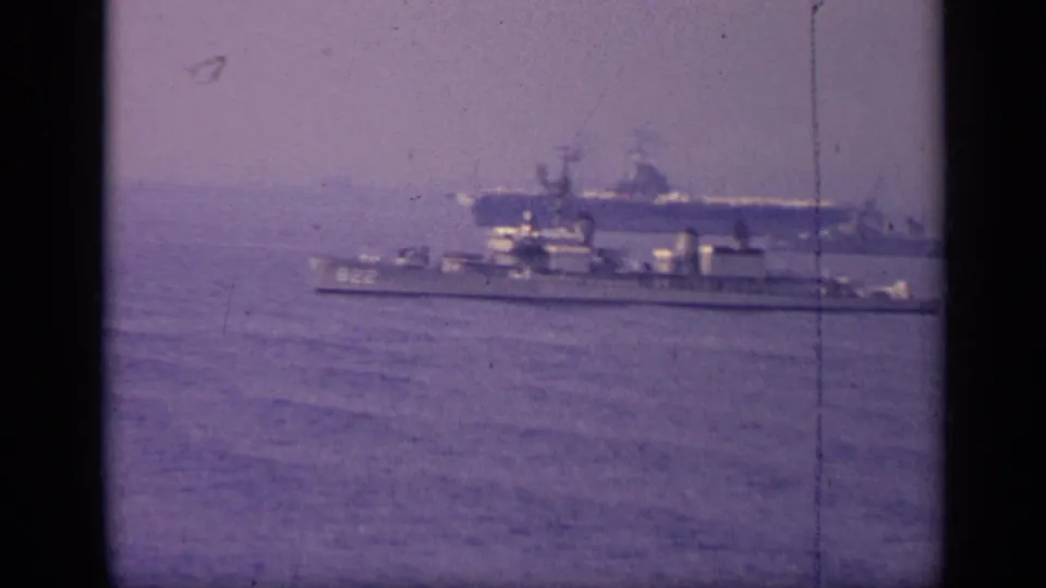 1962 Italy 8mm frame showing a carrier-like naval vessel beyond a smaller ship marked 822 on blue water.