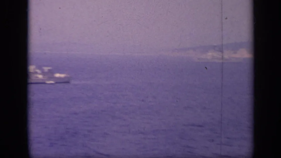 1962 8mm view of a distant vessel moving across calm coastal water in Italy, with land faintly visible behind it.