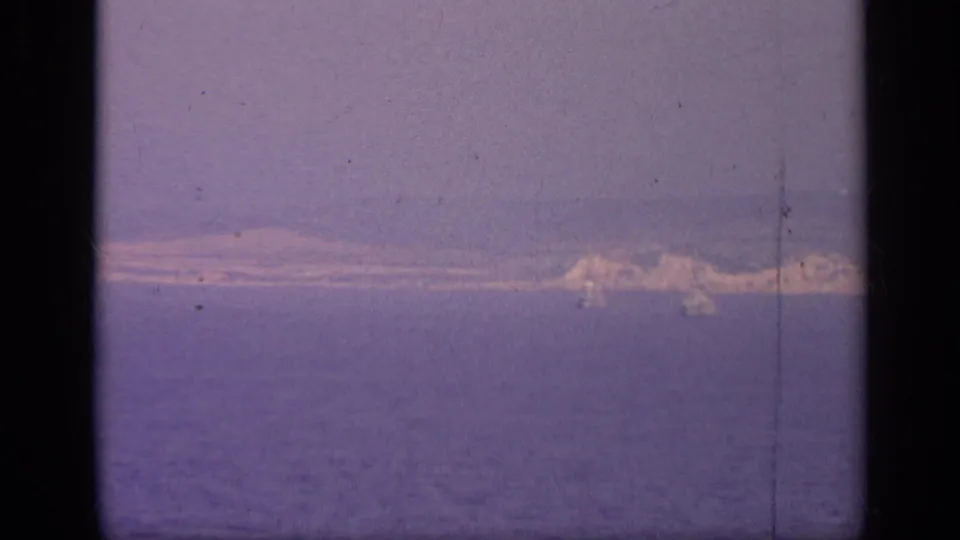 A distant bright vessel or ship structure sits near the right horizon in 1962 Italy coastal footage, softened by grain and color shift.