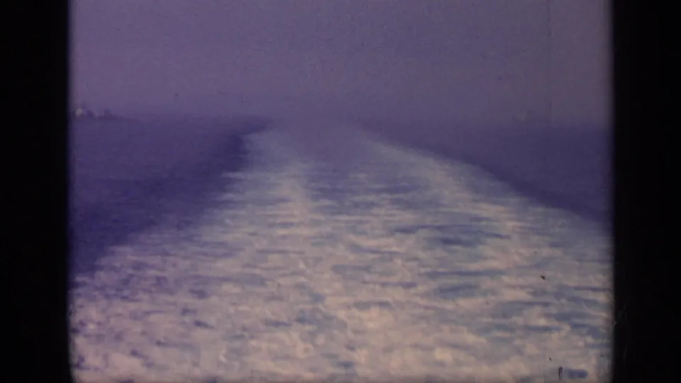 1962 Italy 8mm frame with a long foamy wake crossing textured sea beneath a hazy purple-blue horizon.