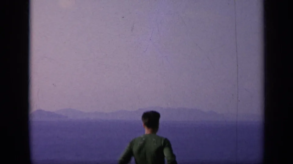 1962 Italy 8mm frame with a man seen from behind on deck, looking over the sea toward faint landforms.