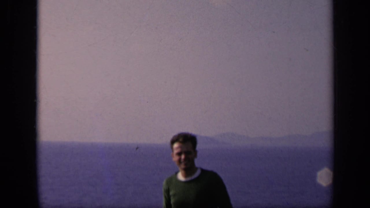 Smiling man in a green sweater stands before water and distant land.