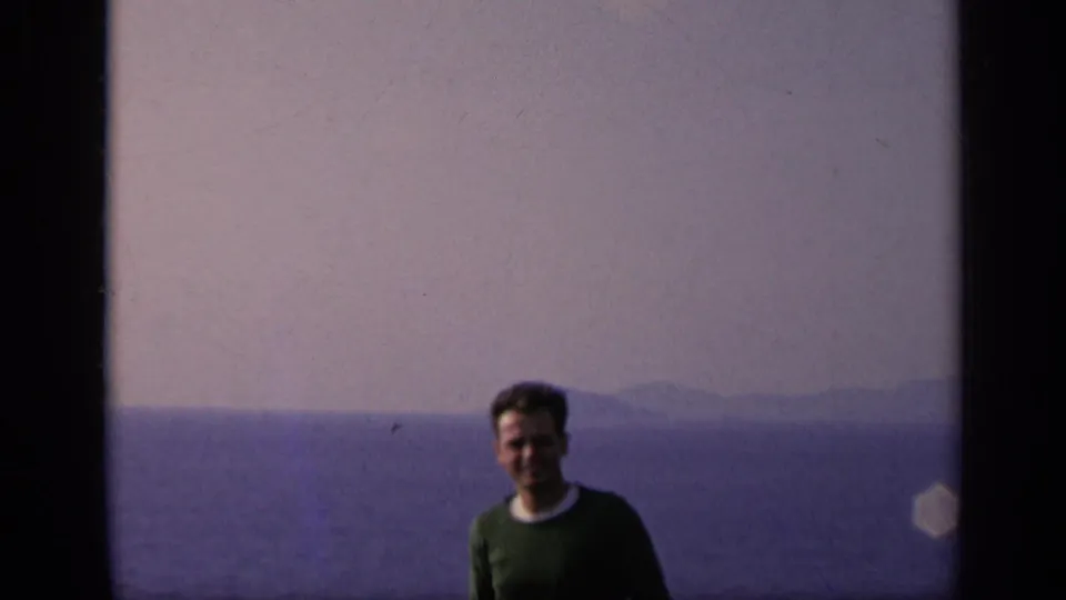 1962 Italy home-movie frame of the smiling man facing the camera with sea and distant shoreline behind him.