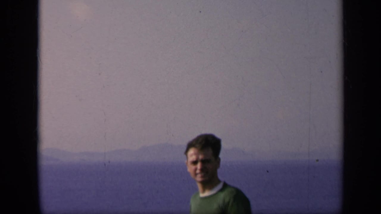 Closer 1962 Italy home-movie view of the man in a green sweater, framed against water and a pale mountain horizon.