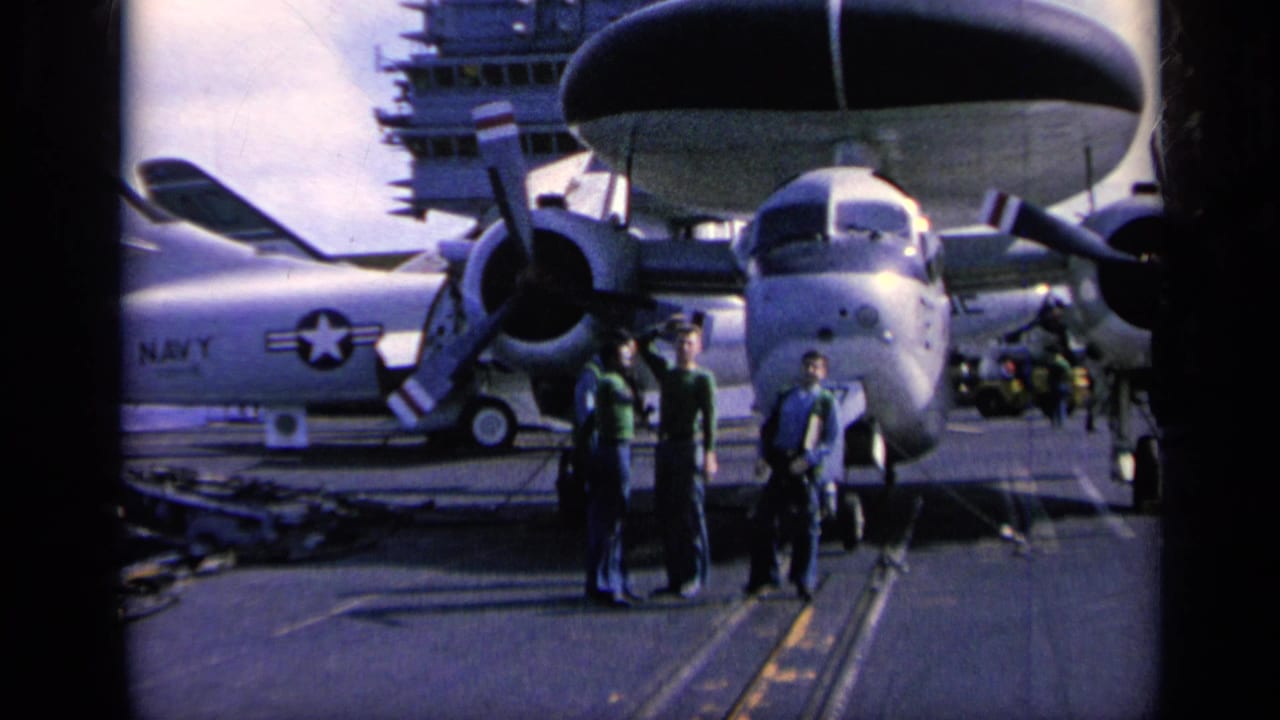 1962 home-movie frame on an aircraft carrier deck, with men near a large Navy propeller aircraft and uniformed sailors nearby.