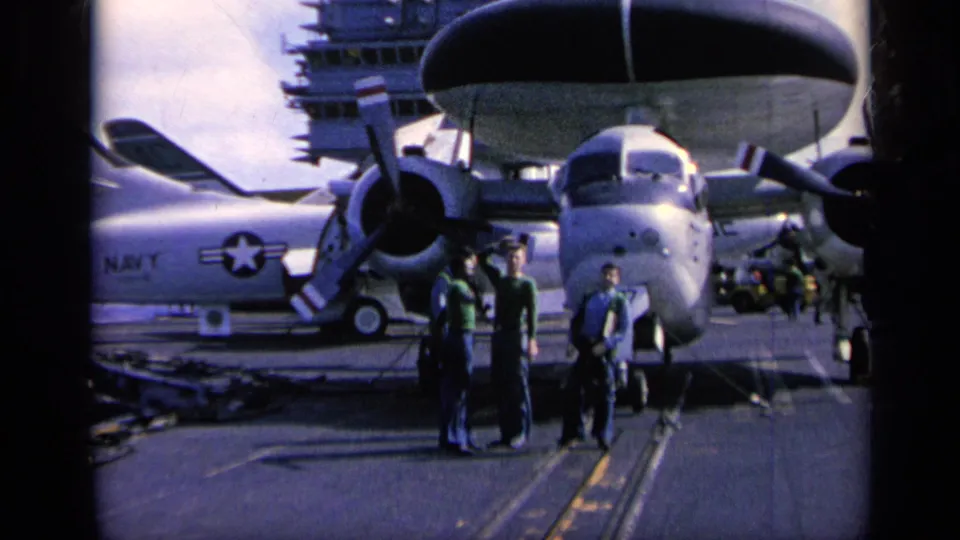 1962 home-movie frame on an aircraft carrier deck, with men near a large Navy propeller aircraft and uniformed sailors nearby.