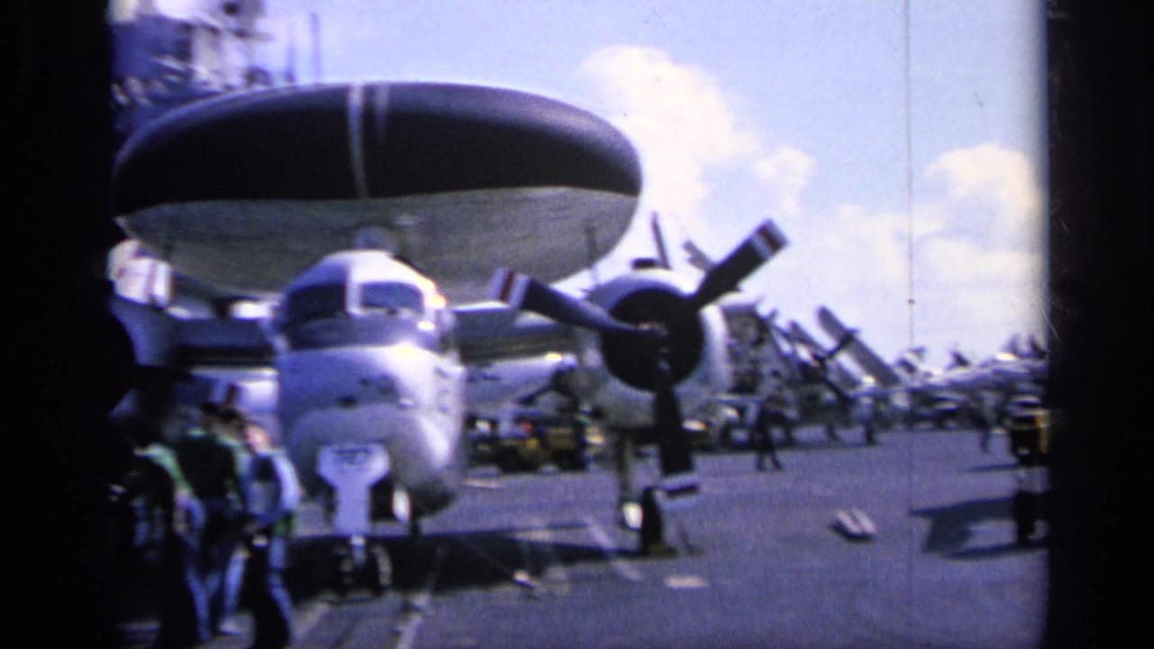Aircraft parked on a carrier deck with people near the nose and a large radome above.