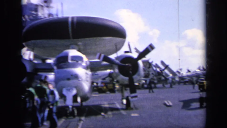 1962 8mm view of a carrier flight deck, with a propeller aircraft and large radome in front and people standing nearby.