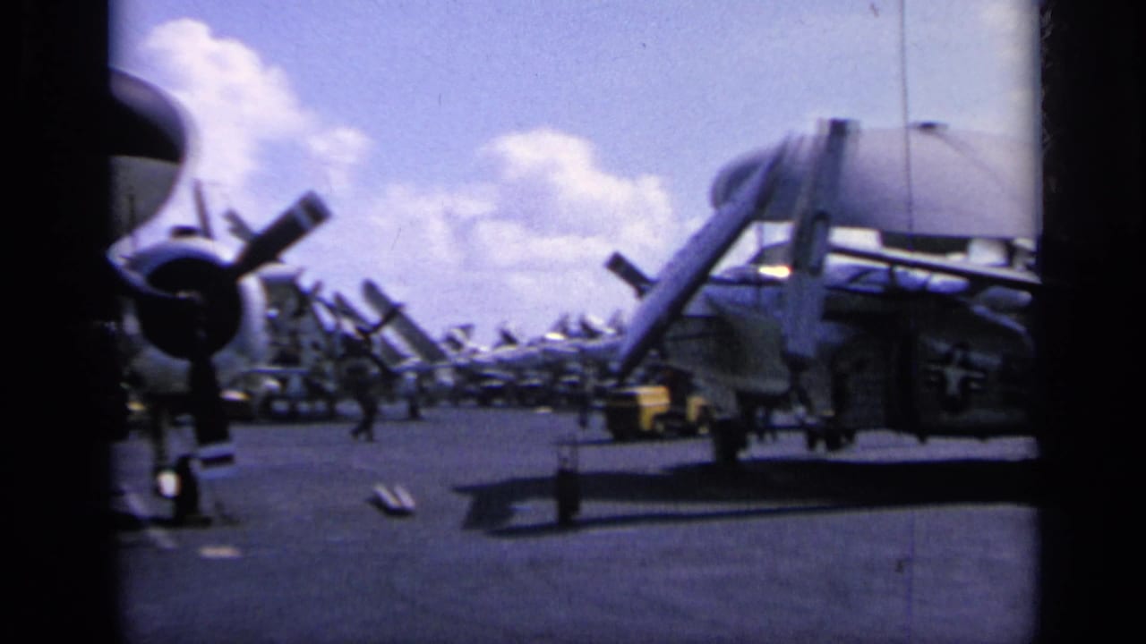 Wide 1962 carrier-deck view showing rows of parked propeller aircraft, a partial aircraft wing or boom at right, and crew figures in the distance.