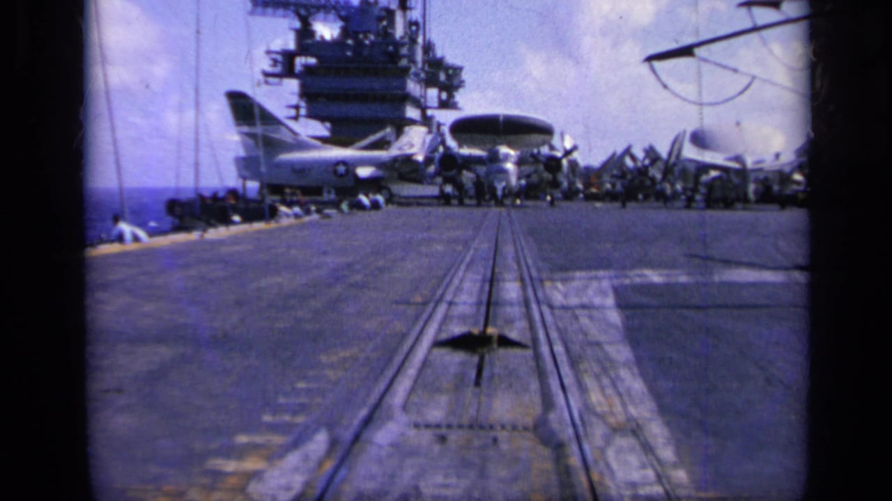 Handheld 8mm view down the carrier deck, with metal deck tracks leading toward the island structure, parked planes, and ocean at left.