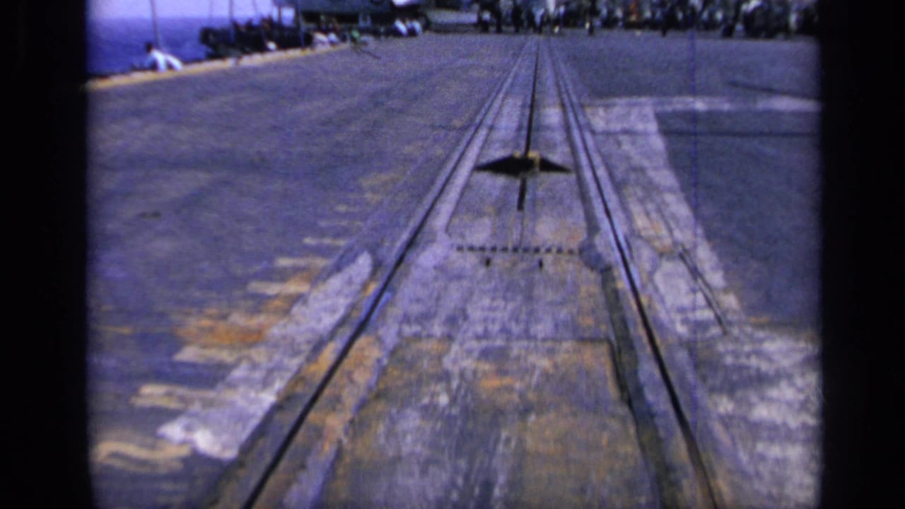 Low 1962 8mm view along the carrier deck surface, showing tracks, weathered markings, and a line of distant people near parked aircraft.