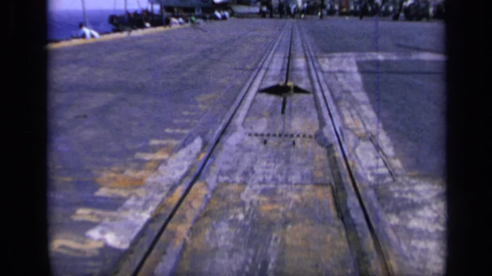 Low 1962 8mm view along the carrier deck surface, showing tracks, weathered markings, and a line of distant people near parked aircraft.