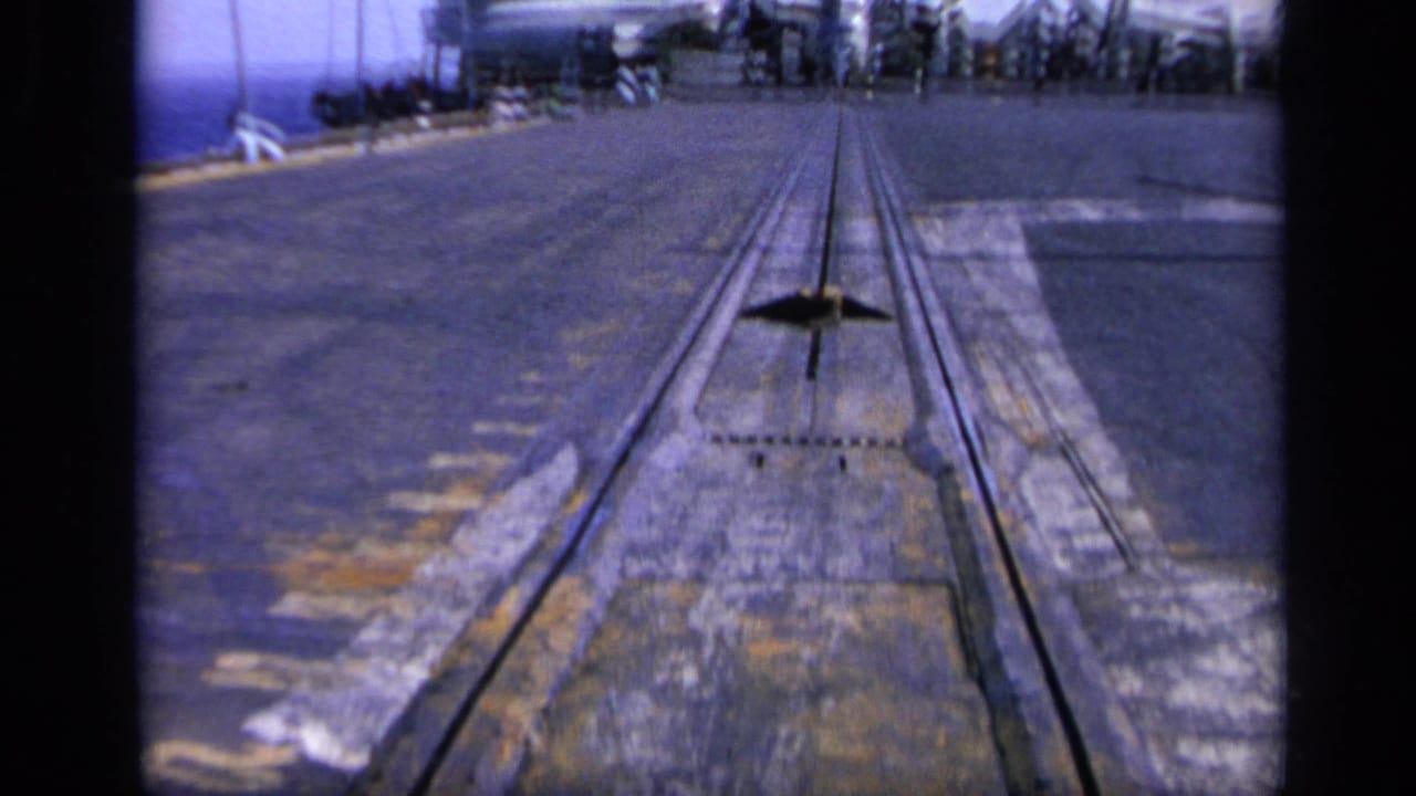 Low view along an aircraft carrier flight deck with rails and weathered surface markings.