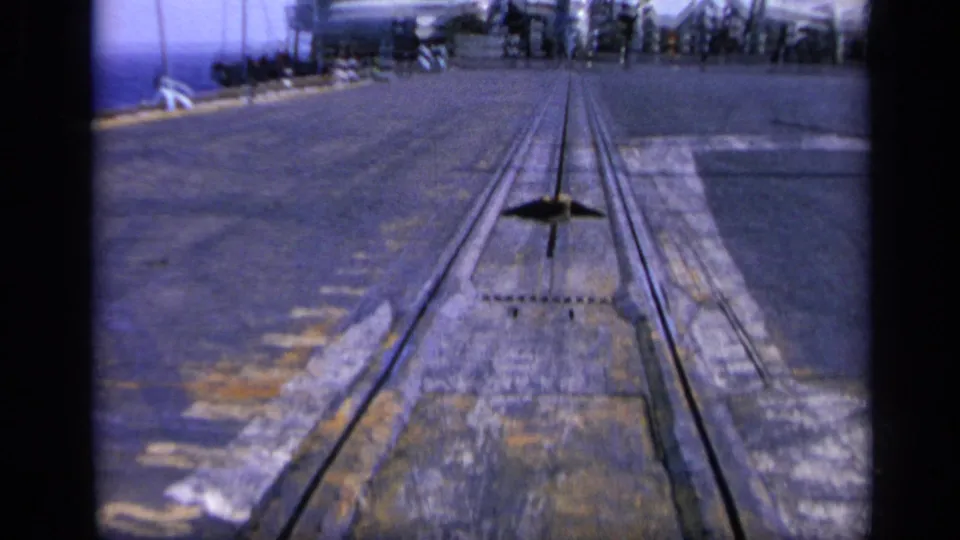 1962 8mm frame from Italy showing a low view along a large aircraft carrier flight deck with rails leading toward distant aircraft and the island.