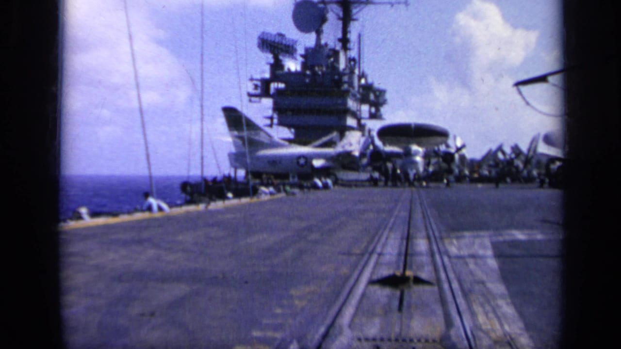 1962 8mm frame from Italy showing an aircraft carrier deck at sea with the island, radar equipment, and parked aircraft under a bright sky.