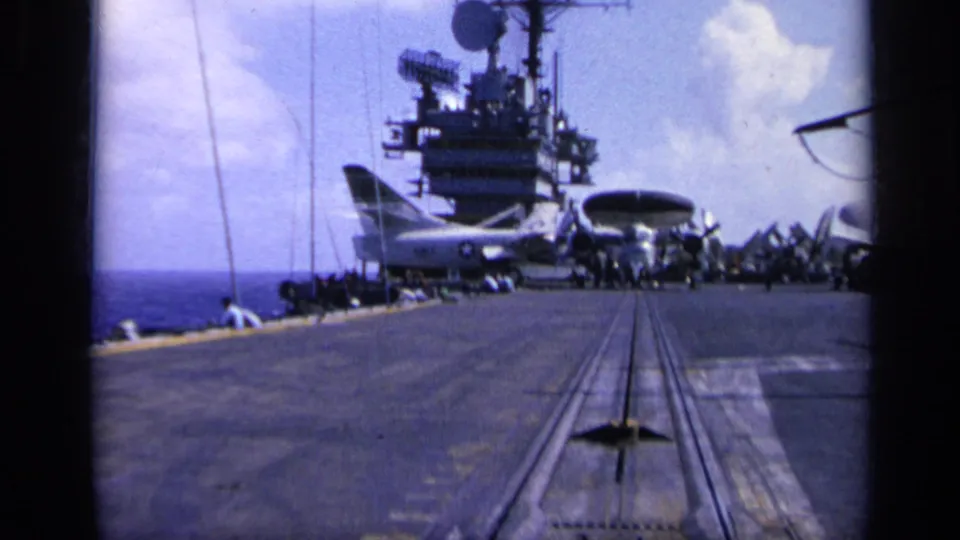 1962 8mm frame from Italy showing an aircraft carrier deck at sea with the island, radar equipment, and parked aircraft under a bright sky.