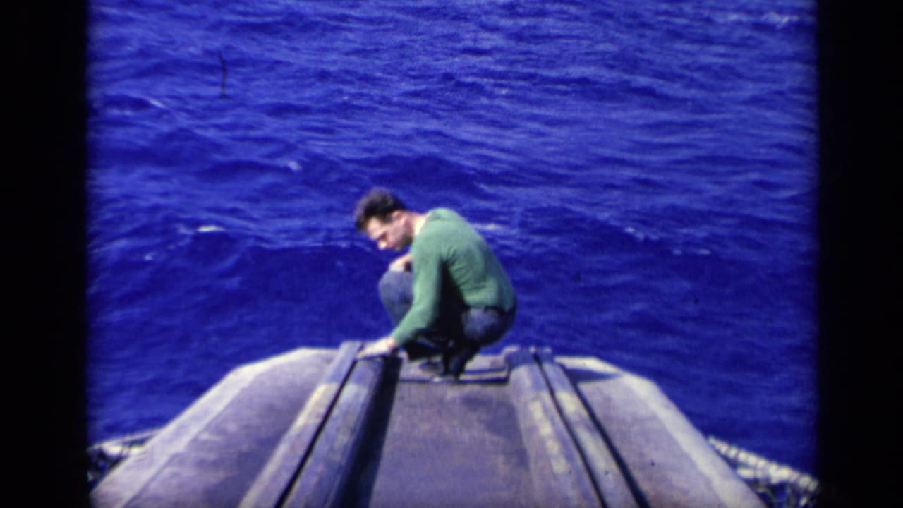 1962 8mm frame from Italy showing a man in a green long-sleeved shirt crouched at the carrier edge above the deep blue sea.