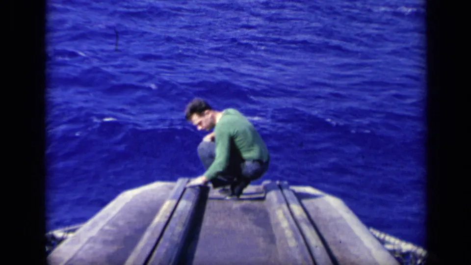 1962 8mm frame from Italy showing a man in a green long-sleeved shirt crouched at the carrier edge above the deep blue sea.