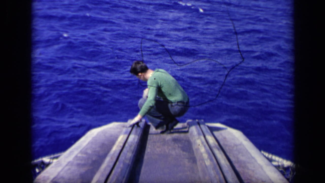 1962 8mm frame from Italy showing the man still crouched at the carrier edge, with open blue ocean behind and below him.