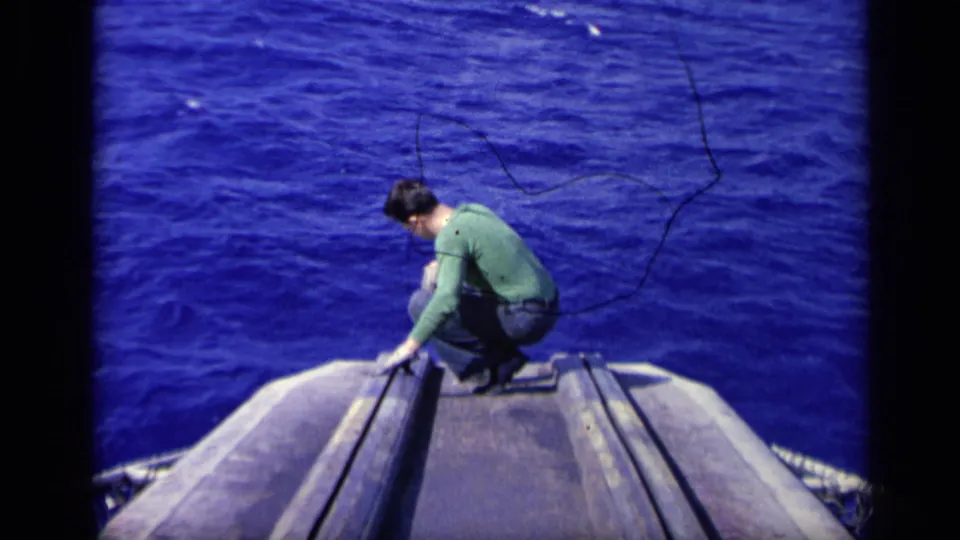 1962 8mm frame from Italy showing the man still crouched at the carrier edge, with open blue ocean behind and below him.
