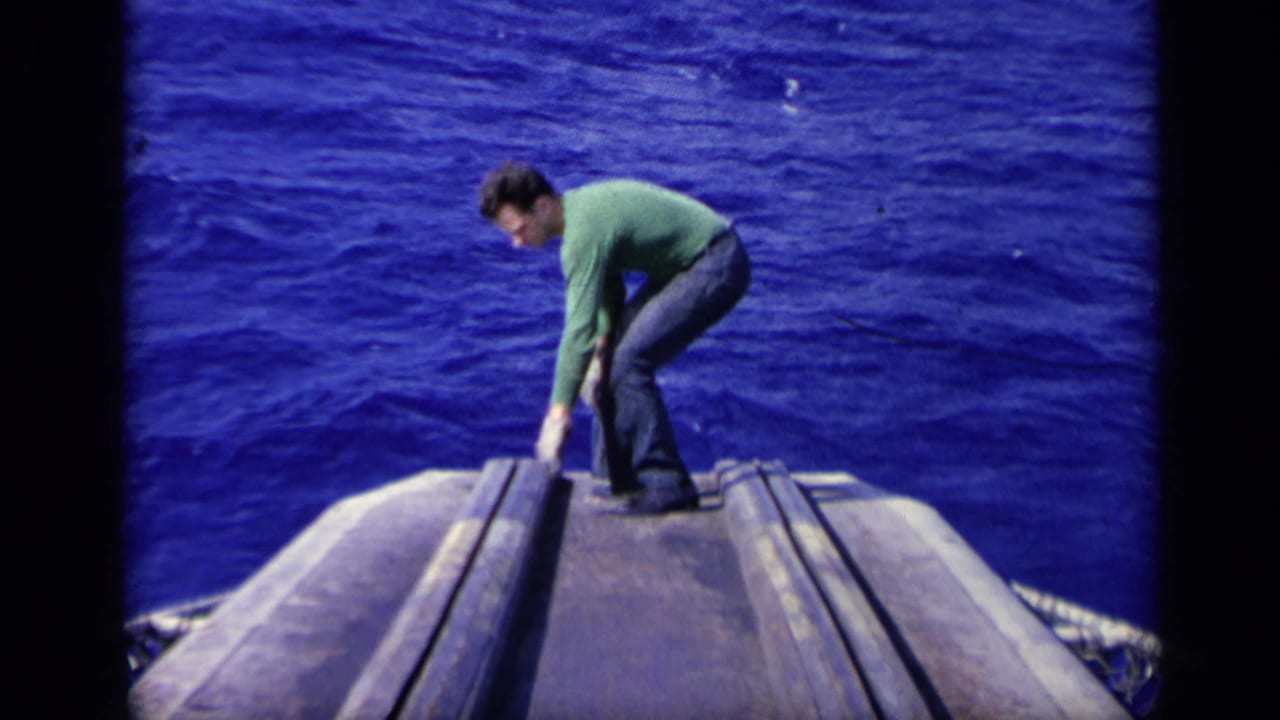 Man in a green sweater crouches on a ship deck with open blue water behind him.