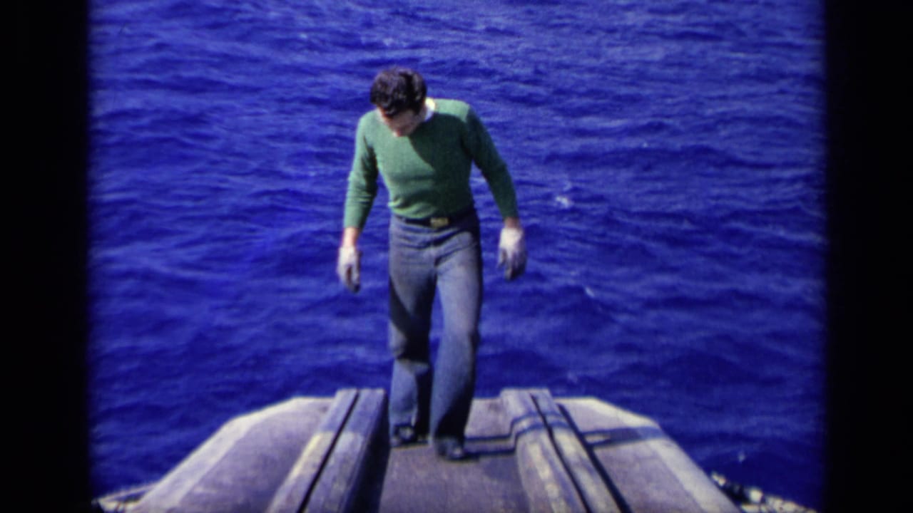 The man stands on the deck of a vessel at sea, looking down as blue ocean surrounds the frame in 8mm footage.