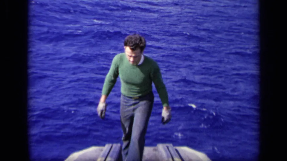 A man in a green sweater walks toward the camera along a vessel deck, with open ocean behind him in 1962 8mm footage.