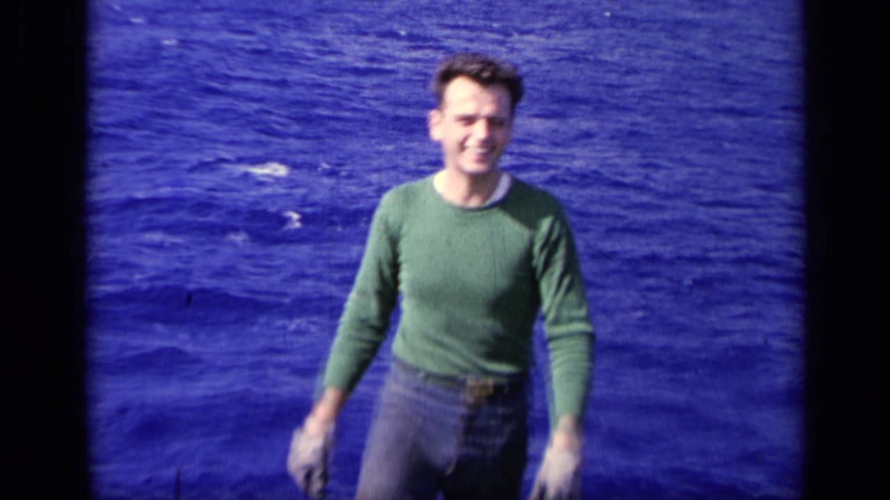 A smiling man approaches the camera on a ship deck, framed closely against saturated blue water in 1962 8mm home-movie footage.