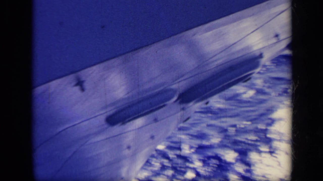 Blue-cast 8mm frame of a boat surface cutting past white ocean spray, with visible grain and dark frame borders.