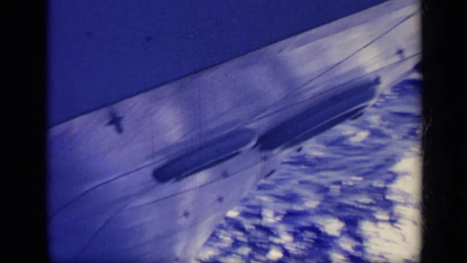 Blue-cast 8mm frame of a boat surface cutting past white ocean spray, with visible grain and dark frame borders.